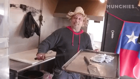 Video gif. Man in a kitchen wearing a frayed straw hat and holding a sheet tray, which he taps with the sword he's holding, waving it as us as he smiles and dances.