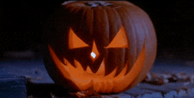 Movie gif. A jack-o-lantern in Halloween 6 lights up with a flickering candle inside. It is a mean-looking smile of sharp teeth, sitting on a porch as wind blows leaves behind it.