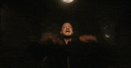 Music video gif. From the video for Imagine Dragon's Eyes Closed, low angle circling around Dan Reynolds pumping his arms in the air pinkies extended, in a tiled room with strobe lighting.