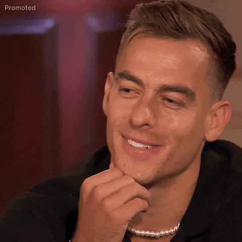 Sponsored gif. Aaron Erb, a contestant from season 21 of The Bachelorette, rests his hand under his chin with a playful smirk. He smiles and waves his hand with indifference, mouth forming a downturned smile like he's saying, "You tell me."