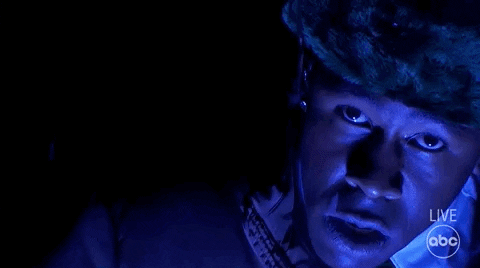 TV gif. From the 2021 American Music Awards, we see a slow zoom out on Tyler the Creator in the dark, staring at us and breathing heavily. He is tinted dark blue, and wears a puffy coat and an ushanka as snow falls around him.