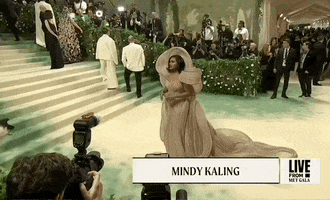 Met Gala 2024 gif. Camera zooms on Mindy Kaling holding her hands delicately in front of her waist as she poses for pictures. She's wearing a champagne-colored form-fitting Gaurav Gupta gown with dramatic loops of fabric that sweep out and around her shoulders forming a giant loose knot at her upper back.