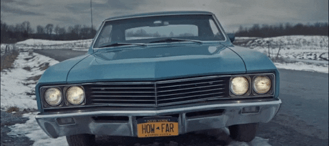 Video gif. Shot of the front of a blue classic American car with a license plate that reads, "How Far."