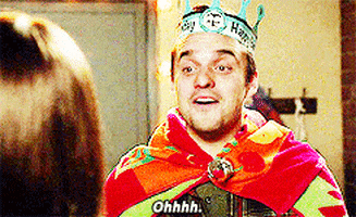 TV gif. Jake Johnson as Nick Miller on New Girl wears a paper crown and a towel as a cape looks at someone with a smile and says, “Ohhhh.”