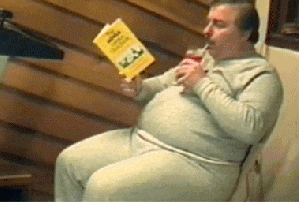 Video gif. A heavyset man in gray sweats relaxes in a wooden chair, reading a book while sipping a drink through a straw. We tilt down to see that his legs are "running" on a treadmill.