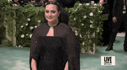 Met Gala 2024 gif. Lily Gladstone smiles with tight lips as we zoom out to see her sparkly black Gabriela Hearst cape and gown.