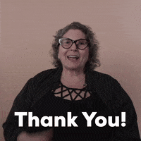 Reaction gif. A Disabled white woman with kinky curly gray hair and big wine-colored cat-eye glasses says "thank you," in ASL and English, punctuating by clutching her heart sweetly.