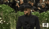 Met Gala 2024 gif. Closeup of the top of Bad Bunny's all navy-washed Margiela smoking jacket with decorative white stitching worn over a black satin corset. He's wearing an oversized puffy beret with floral details and diamond-shaped dark gold-framed sunglasses.