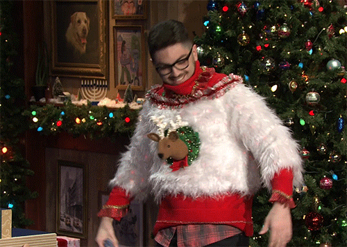 Tonight Show gif. Man stands on a stage decorated to look like an old house during Christmas time. He proudly shows off his ugly sweater which has a foam head of a reindeer coming off of it and is made out of a furry white material.
