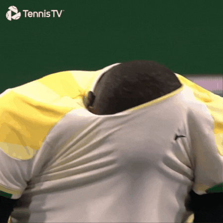 Sports gif. Frances Tiafoe, an American tennis player, is on the court and has his head in his shirt. His head pops back out and he grimaces at us. 