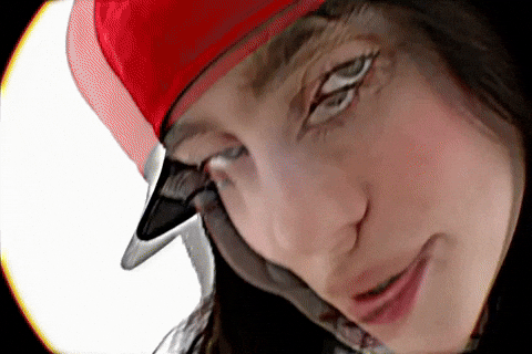Music Video gif. From Billie Eilish's music video for "LUNCH", Billie is on a stark white background dressed in a long sleeve white shirt with a yellow and black jersey over the top, with a red hat on. She starts very close to the camera singing her song while tilting her head left and right.