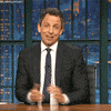 TV gif. Seth Meyers on Late Night with Seth Meyers sits at his desk and gives a big shrug to the audience while a wide smirk spreads across his face. 