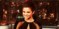 Celebrity gif. Selena Gomez smiles, facing downward as if flattered, at a talk show appearance.
