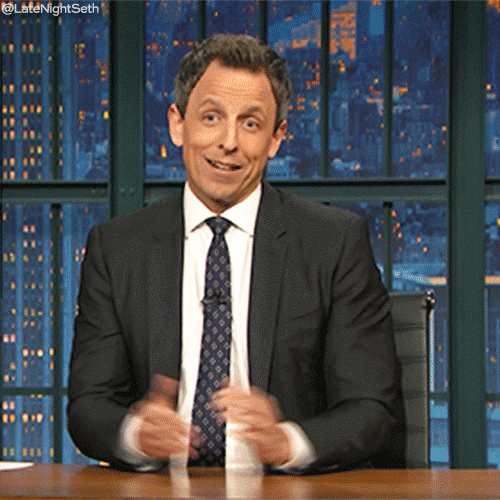 TV gif. Seth Meyers on Late Night with Seth Meyers sits at his desk and gives a big shrug to the audience while a wide smirk spreads across his face. 
