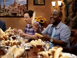 The Tonight Show gif. Jimmy Fallon, Steven Higgins, and the Roots all sit around, being silly, and generally goofing off at a dinner table with wine glasses and so many baskets of breadsticks. Steve Higgins has shoved breadsticks onto each of his fingers and holds a breadstick in his mouth like a cigar. The Roots toss the breadsticks in the air and use them as drumsticks on the table. 