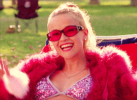 Movie gif. Reese Witherspoon as Elle Woods in Legally Blonde wears a sparkly bikini top and a fuzzy pink jacket while laying on a beach lounge chair. She waves at someone and smiles. We then cut to Elle at her graduation, in cap and gown, smiling just as widely as before.