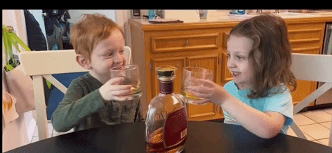 SNL gif. Two toddlers are sitting with cups and a bottle of bourbon. They clink glasses and giggle at each other.