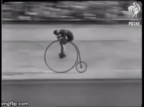 Video gif. An old black and white video of a bicycle race. Men pedal fast along a curved race track on old timey bicycles that have one big wheel in the front.