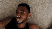 Video gif. A man lies deadpan on the floor. The view spins as it moves farther away from him.