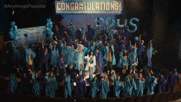 Movie gif. From Anything's Possible, a graduating class in light and dark blue gowns jump for joy and toss up their graduation caps.