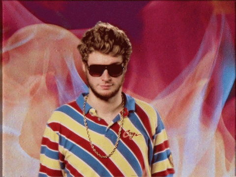 Celebrity gif. Standing in front of a background of flames, Rapper Yung Gravy, raises his hands into the air. 