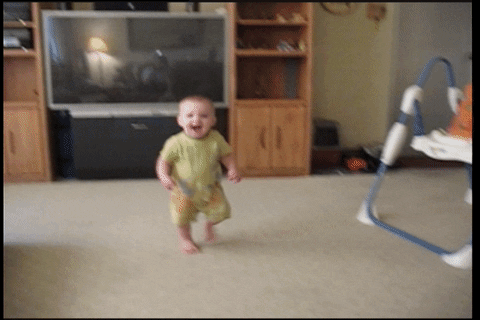 Video gif. Baby happily runs toward us in a living room, then a cat jumps in the frame to attack and knocks the baby to the carpet, where he lays crying.