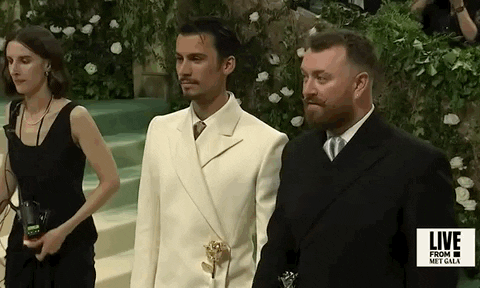 Met Gala 2024 gif. Sam Smith and designer Christian Cowan stand for the cameras wearing matching suits, Sam in black, Christian in ivory.