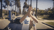 Celebrity gif. Wearing an unbuttoned Dodgers jersey, Rapper Lil Dicky stands in front of a Lamborghini, clapping his hands enthusiastically.