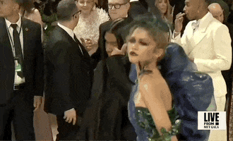 Met Gala 2024 gif. Zendaya wearing a peacock-colored Maison Margiela gown by John Galliano embellished with true-to-life grapes and hummingbirds, poses with a brooding confidence, then struts away to her next pose.