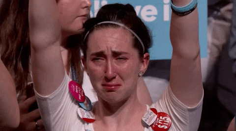 Video gif. Woman wearing many button pins on her shirt holds her hands up in the air as she tries not to sob.