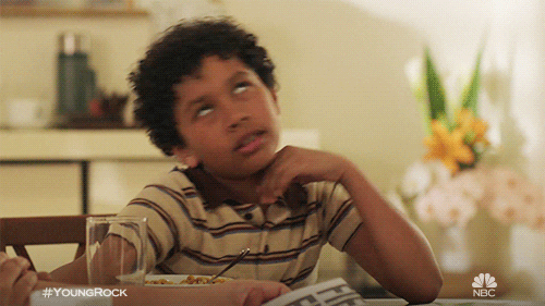 TV gf. Adrian Groulx as young Dwayne Johnson on Young Rock sits at a kitchen table. He has his hand on his cheek and he flops to his side, leaning onto the table, as he rolls his eyes and sighs.