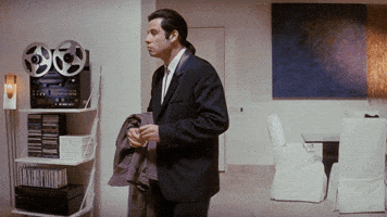 Meme gif. Confused Travolta, standing in a living room, extending his hand and glancing around, puzzled.