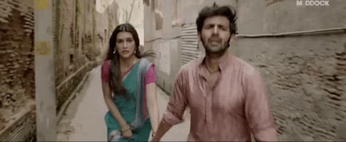Movie gif. Kartik Aaryan as Vinod Shukla runs down an alley, holding hands with Kriti Sanon as Rashmi Trivedi in Luka Chuppi.