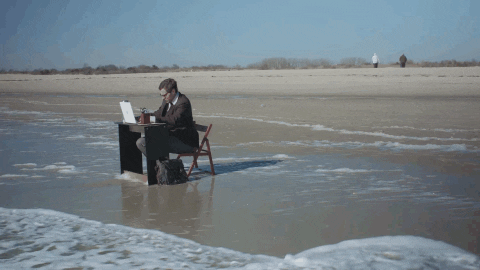 Video gif. Man works at a desk on the shore of a beach, continuing to type even as he stands up when the tide rolls in.