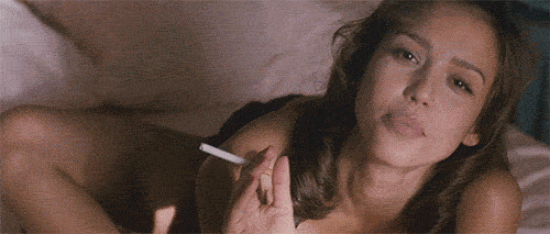 Celebrity gif. Holding a cigarette, Jessica Alba seductively rolls onto her belly in bed and smiles at us.