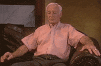 Video gif. An old man sits up straighter on a couch and waves his arms up and down like he is doing jumping jacks while seated.
