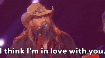 ACM Awards gif. Man in tan cowboy hat with guitar strapped around him sings "I think I'm in love with you" into mic stand.