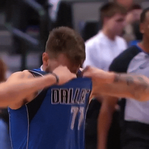 Sports gif. Luka Doncic of the Dallas Mavericks rips his jersey collar during a game in frustration as he walks with his head down across the court.