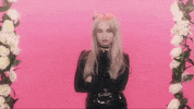 Celebrity gif. Kim Petras stands with her arms crossed in front of a vibrant pink backdrop with white floral decorations on each side. She is completely over it, rolling her eyes dramatically and looking off to the side.