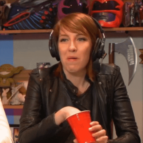 Video gif. Woman in a leather jacket and headphones sits in front of a collection of gamer items. She snacks out of a red solo cup and raises her hand while her eyes widen excitedly.