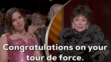 Oscars 2024 GIF. Split screen of Rita Moreno, who is on stage, addressing America Ferrera, who sits in the audience. Moreno congratulates Ferrera, saying "Congratulations on your tour de force." Moreno wears a dark dress with a big ruffled neckline and Ferrera wears a bright pink glittered dress.  Ferrera is touched by Moreno's words and puts a hand on her chest emphatically.