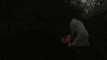 Video gif. Person in a white lab coat launches a ball of flaming hay out of a container in slow motion.