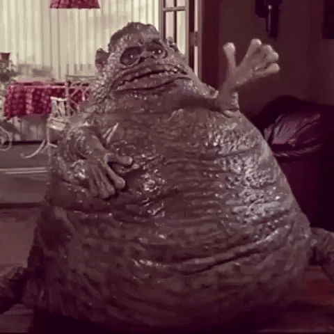 Movie gif. From Weird Science, gigantic shiny Jabba-the-Hutt-type creature in a living room, waving hello.