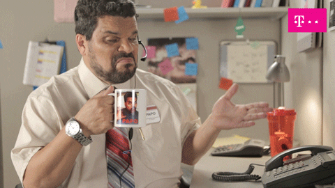 Video gif. Office worker sits at his desk with his headset on and holds a mug with a picture of his face on it. His mouth is turned down at the corners as he shrugs and looks at us up and down. A T-Mobile logo rests in the top right corner.