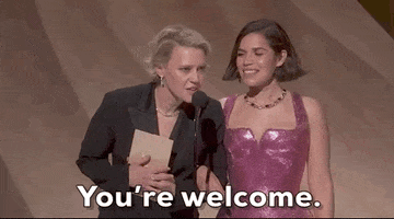 Oscars 2024 GIF. Kate McKinnon and America Ferrera stand on stage and McKinnon is leaned into the microphone, staring intensely without blinking, and says, "You're welcome." 