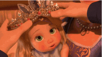 Disney gif. Baby Rapunzel from Tangled has an oversized tiara placed on her head and giggles.