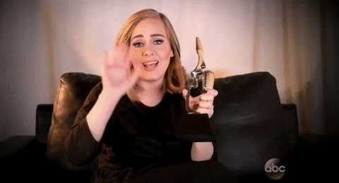 Celebrity gif. Sitting on a sofa, Adele holds up a trophy in her hand and waves hello with her other hand, smiling.