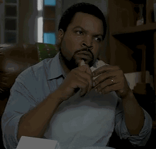Celebrity gif. Ice Cube sits looking skeptical, chewing slowly on the sandwich that he is holding.