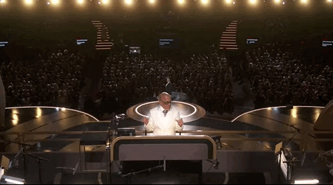 Oscars 2024 GIF. The camera shoots Rickey Minor, symphony conductor of the Oscars, from the stage angle and we see the audience behind him. He's wearing a white suit and he's grooving along to the music he conducts.