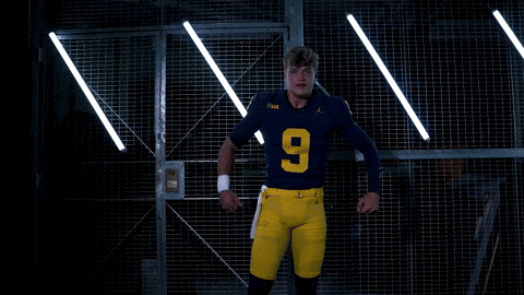 Sports gif. JJ McCarthy, a quarterback for the Michigan Wolverines football team, is wearing his full uniform and he pounds his chest while yelling and coming towards the camera. 